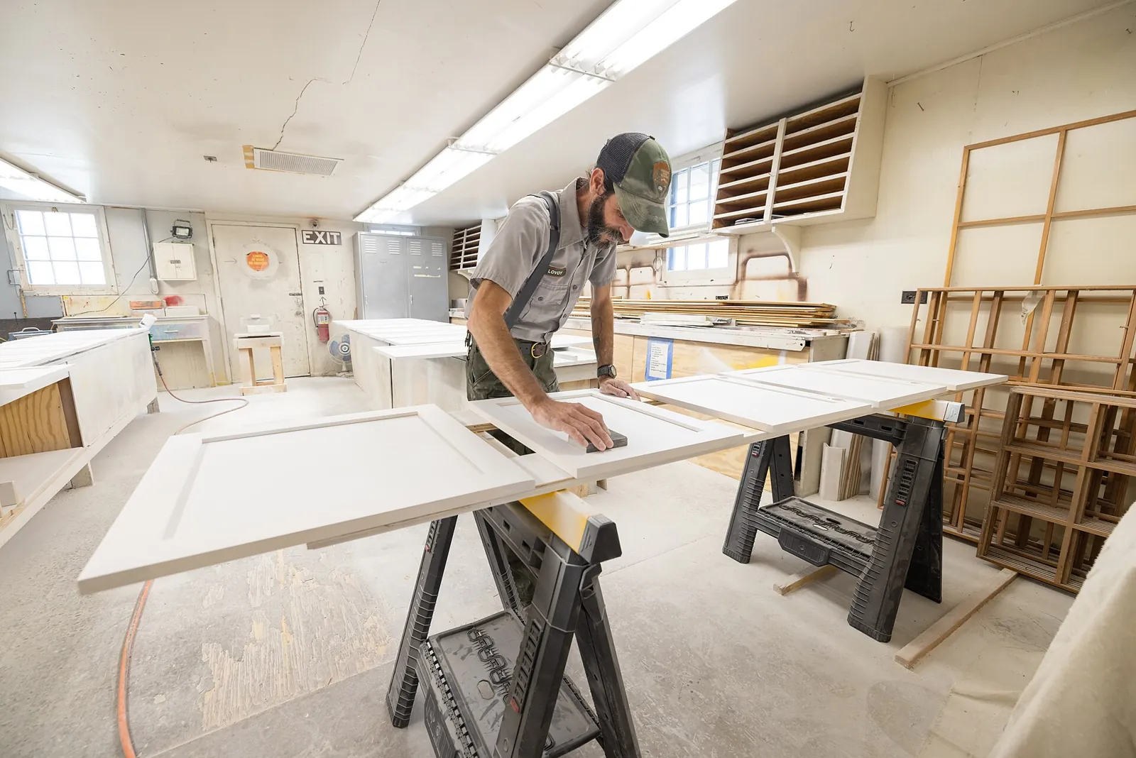 Woodworker hand-sanding a white cabinet door with a sanding block, more doors laid across sawhorses